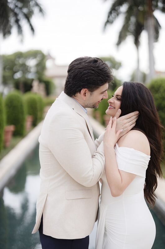 Iviani and Joey face to face beside a palm-lined reflecting pool, his hand on her cheek