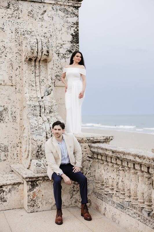 Iviani standing on a coral-stone wall as Joey sits below, the ocean behind them